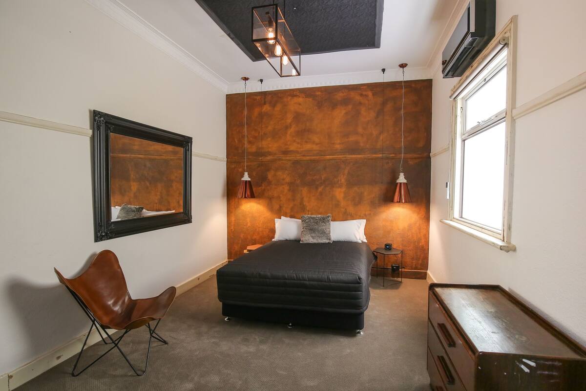 A bedroom with a large bed, a chair, a dresser, a mirror, and a wall-mounted light fixture.