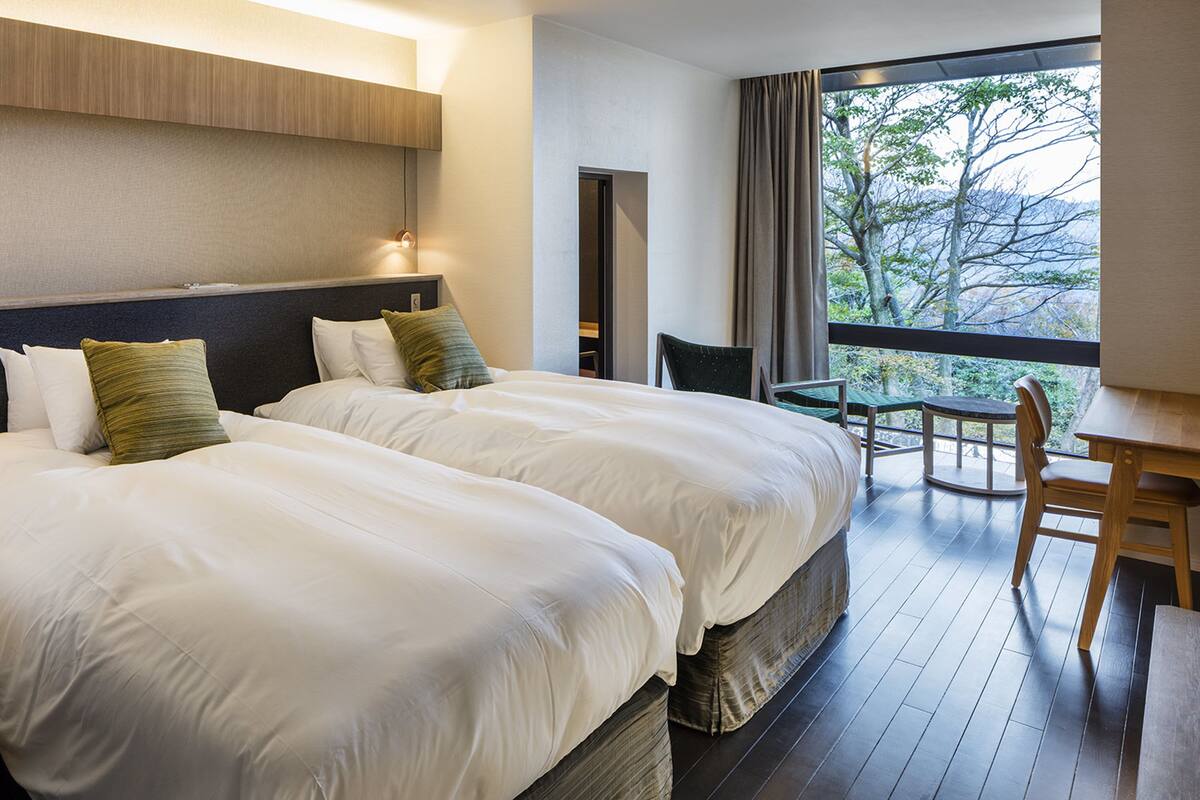 Ein Hotelzimmer mit zwei grossen Betten, einem kleinen Tisch und einem Sessel. Durch ein grosses Fenster ist eine Baumlandschaft zu sehen.