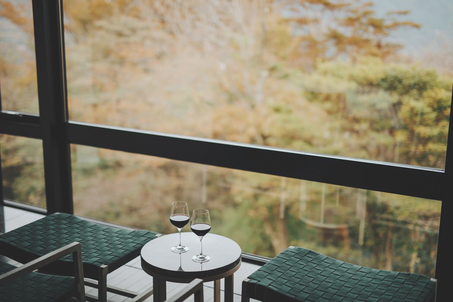 A table with two wine glasses and a view of autumn trees through large windows.