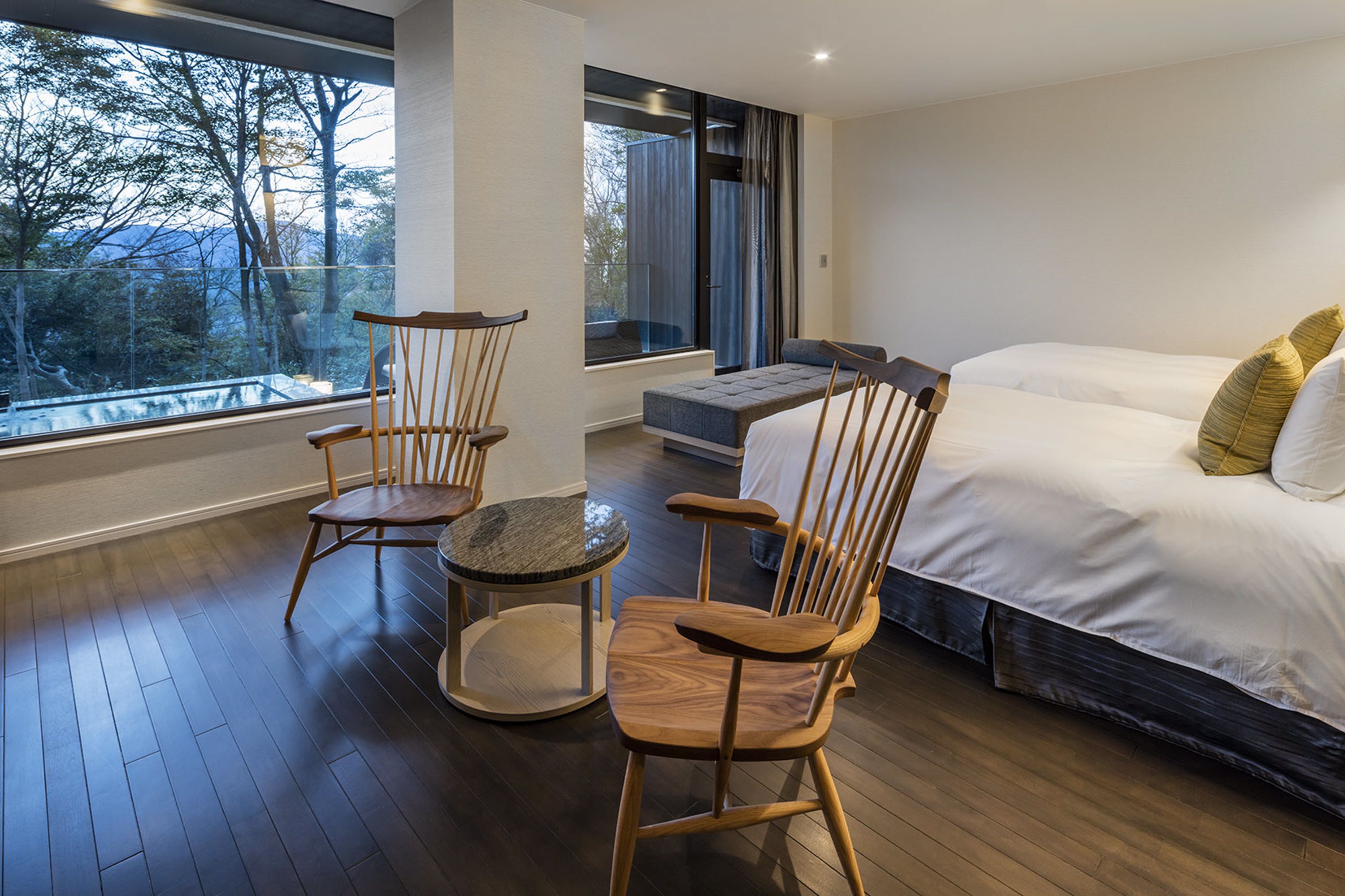 Un dormitorio moderno con una cama grande, dos sillas de madera, una mesita redonda y vistas a árboles a través de un ventanal amplio.