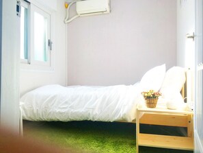 A small, single-bed room with a white bed, a wooden nightstand, a basket of flowers, and a window with a view.