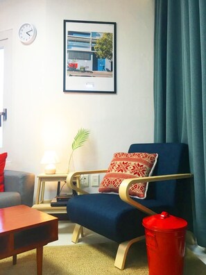 A modern living room with a blue armchair, a small table, a red trash bin, a clock, and a framed picture on the wall.
