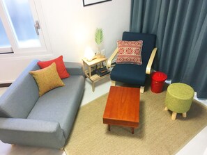 A living room with a grey sofa, a wooden coffee table, a blue armchair, a small side table with a lamp, a green ottoman, and a red bucket.
