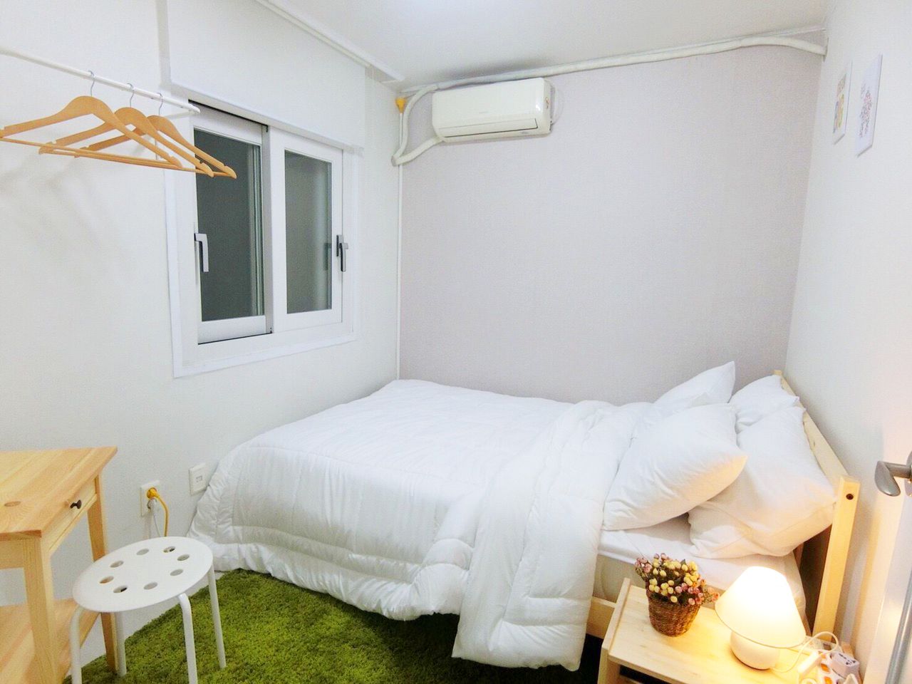 A small, neatly arranged bedroom with a bed, a bedside table with a lamp, a small stool, a window, and a wall-mounted air conditioner.
