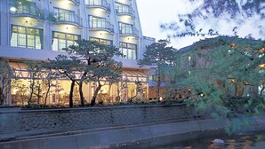 Un hôtel moderne avec une façade en pierre, de nombreux balcons et un élément décoratif aquatique réfléchissant à l’avant.