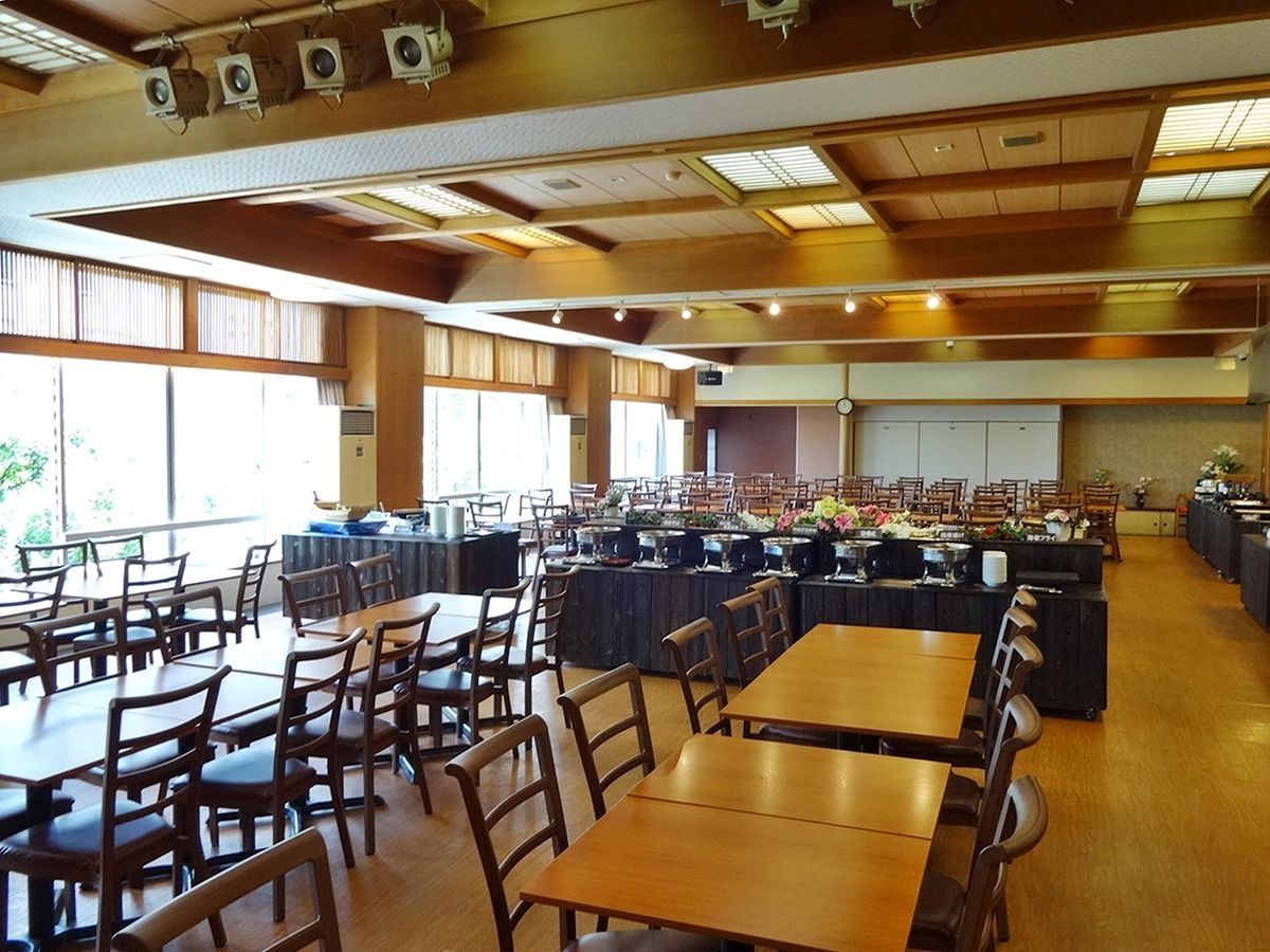 A spacious cafeteria with multiple tables and chairs, large windows, and ceiling lights.