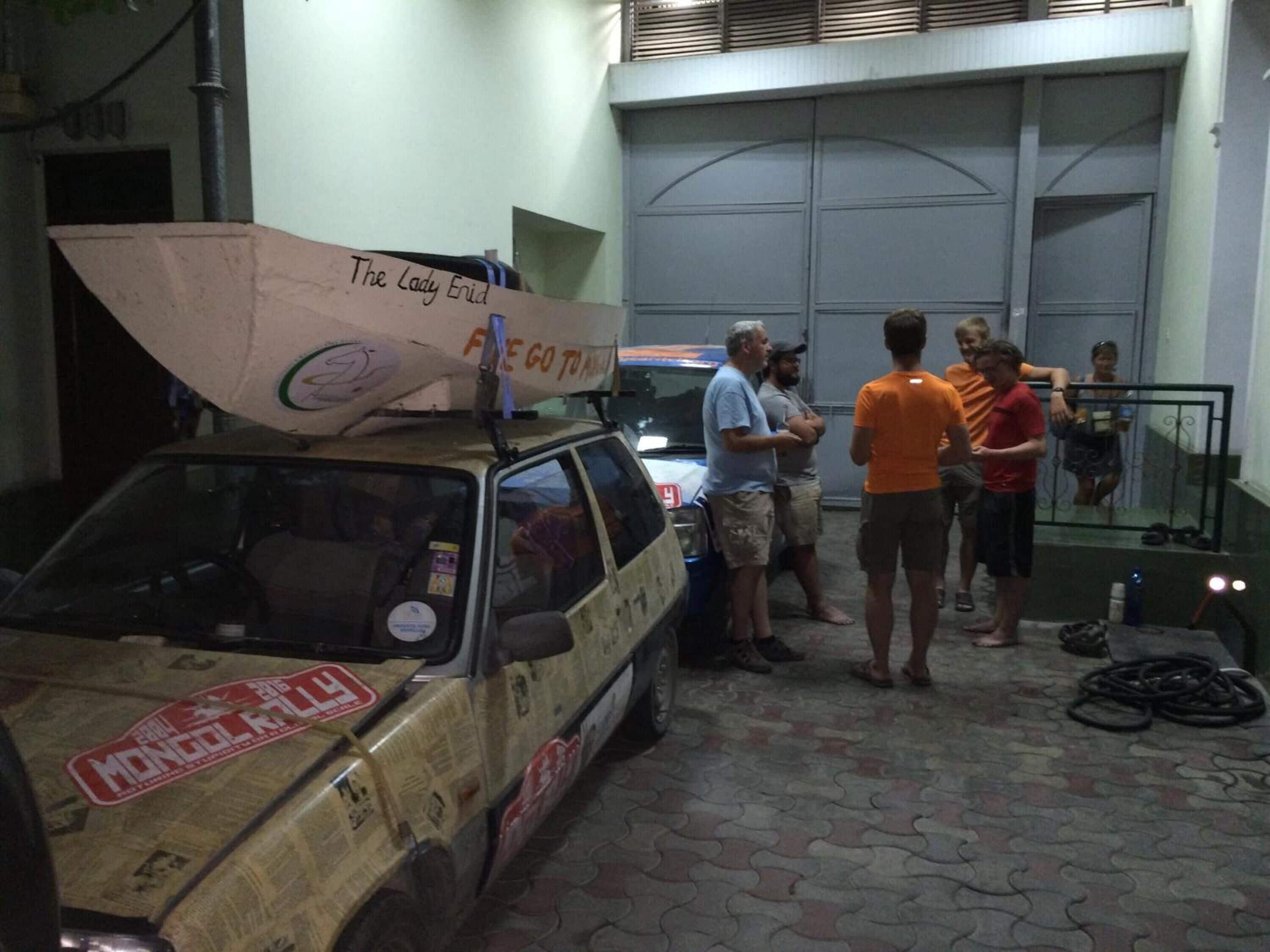A vehicle with a large surfboard on top, parked in a garage-like setting with people standing around.