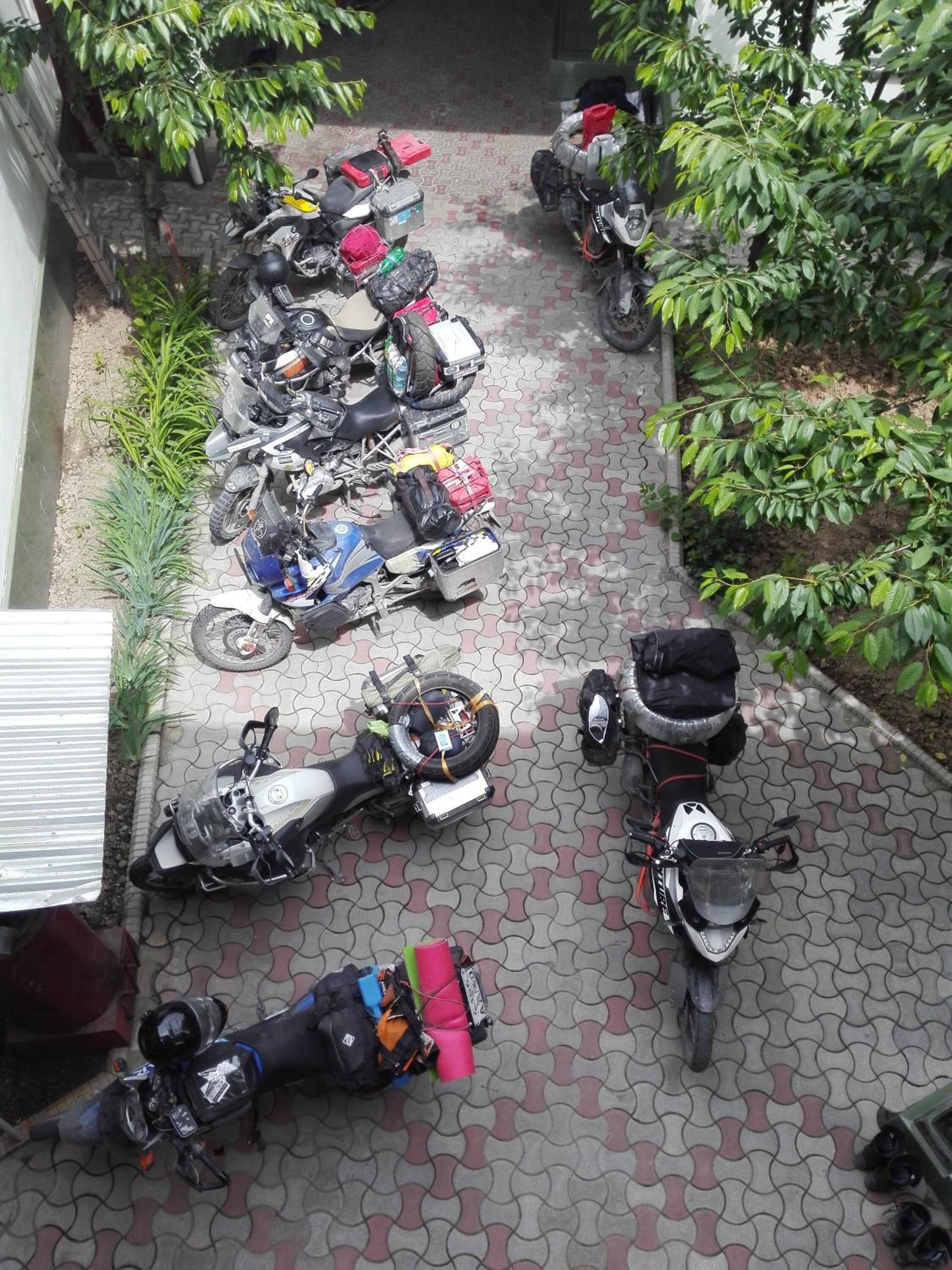 A paved courtyard with multiple parked scooters and motorcycles, surrounded by greenery.