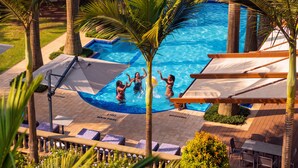 Zona de piscina con sillas de descanso y sombrillas, rodeada de palmeras y una terraza de madera.