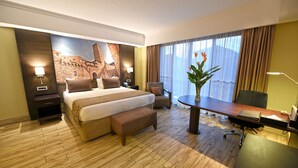 A hotel room with a large bed, a desk, a chair, a small table, and a large window with curtains. - Mestil Hotels & Residences (Kampala)