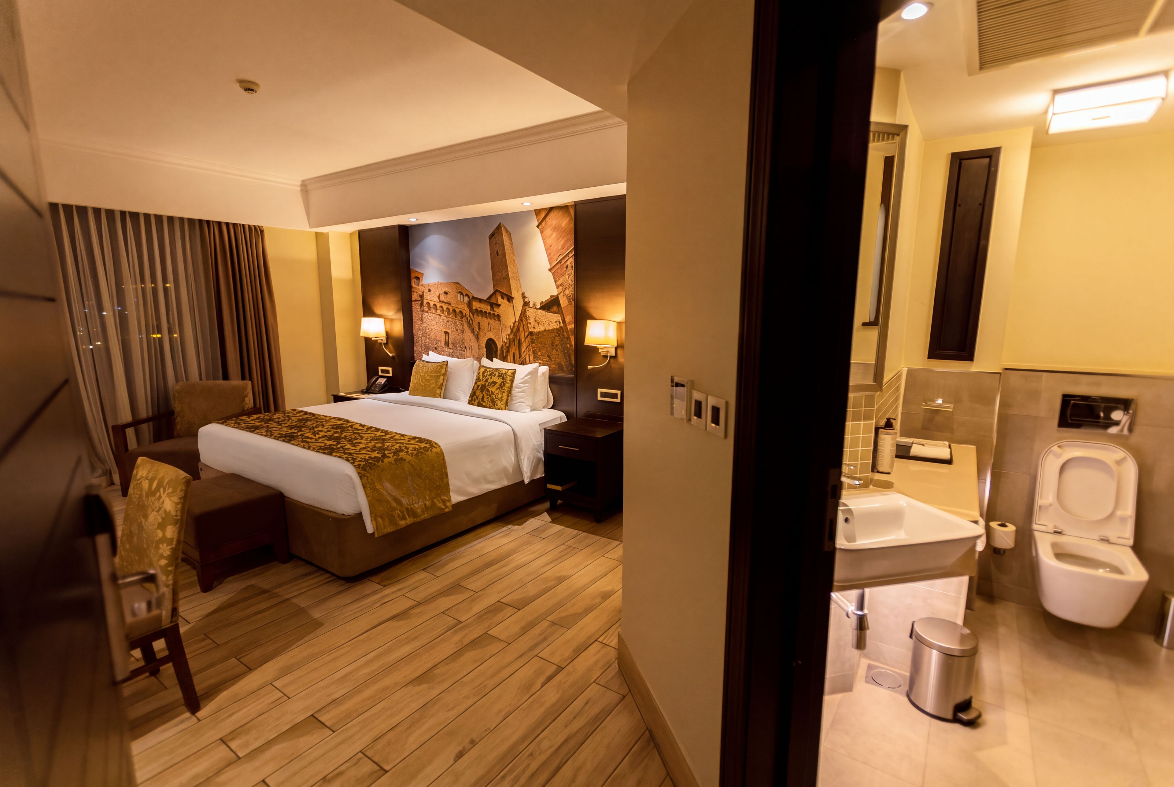 A hotel room with a large bed, a desk, a chair, a bathroom with a toilet and sink, and a mural on the wall.