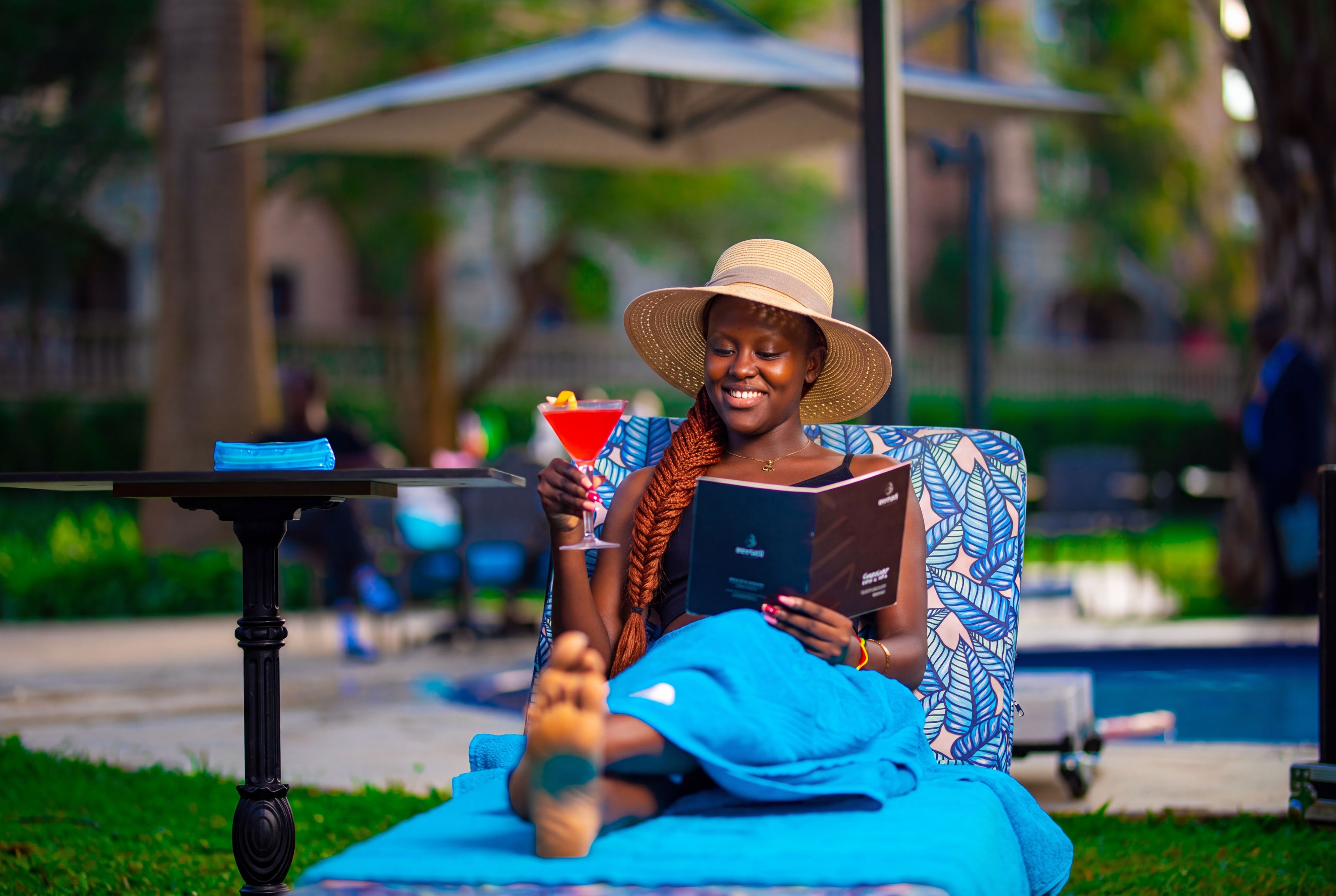 Une personne assise sur un tapis bleu, au bord d’une piscine, avec une boisson et un livre.