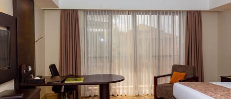 A hotel room with a bed, a desk, a chair, a TV, and a large window with curtains.