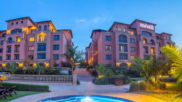 Un hotel con fachada simétrica, varios balcones y un jardín bien cuidado con palmeras y una piscina.