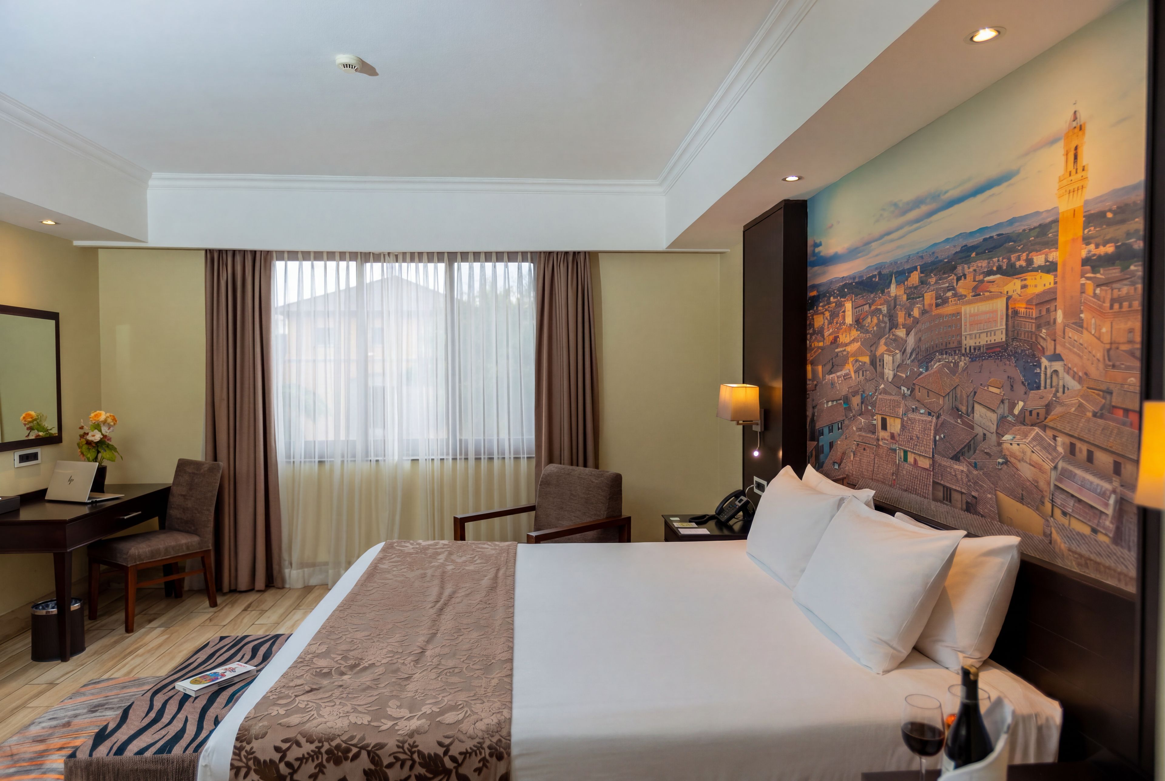A hotel room with a bed, a desk, a chair, a lamp, and a painting of a cityscape.