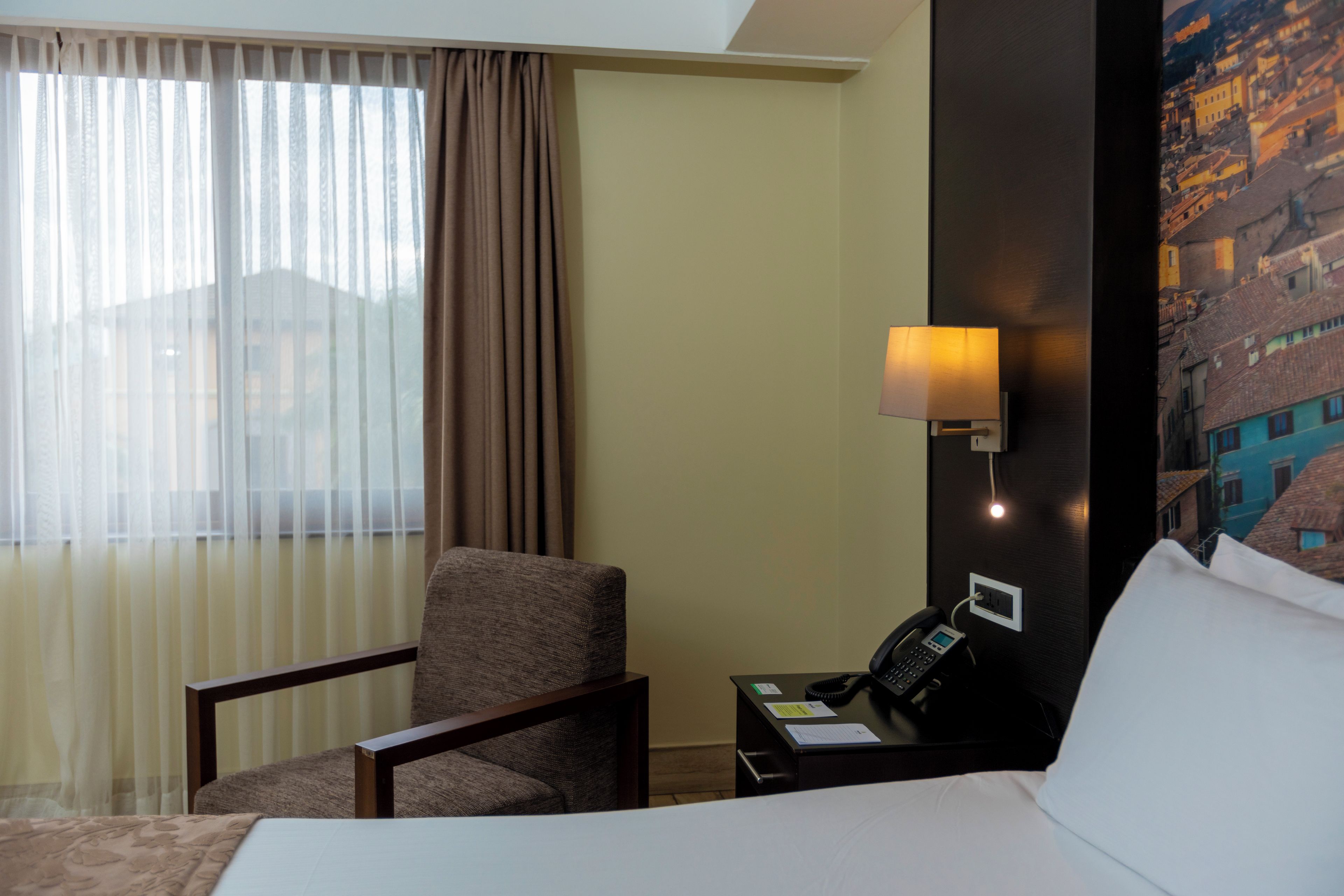 A hotel room with a bed, a chair, a desk with a phone, and a wall-mounted lamp.