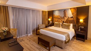 Habitación de hotel con una cama grande, un escritorio, una silla, una mesita con un jarrón de flores y una lámpara de pared.