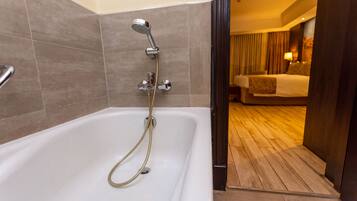 Un baño moderno con bañera, ducha y una habitación visible con cama y cortinas.