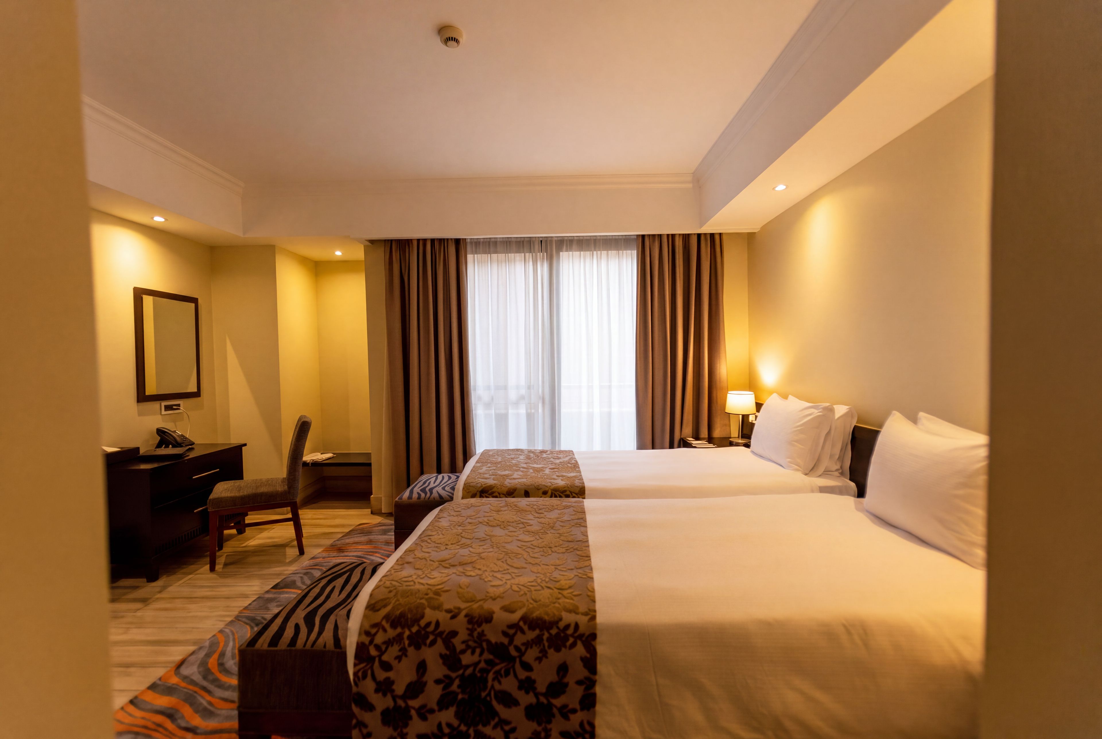 Une chambre d’hôtel avec deux lits, un bureau, une chaise, une lampe et une fenêtre avec des rideaux.