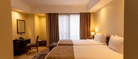 Habitación de hotel con dos camas, un escritorio, una silla, una lámpara y una ventana con cortinas.