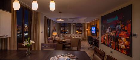 A hotel room with a dining area, a sofa set, a television, and a large painting on the wall.