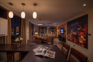 Habitación de hotel con zona de comedor, un conjunto de sofás, televisión y un cuadro grande en la pared.