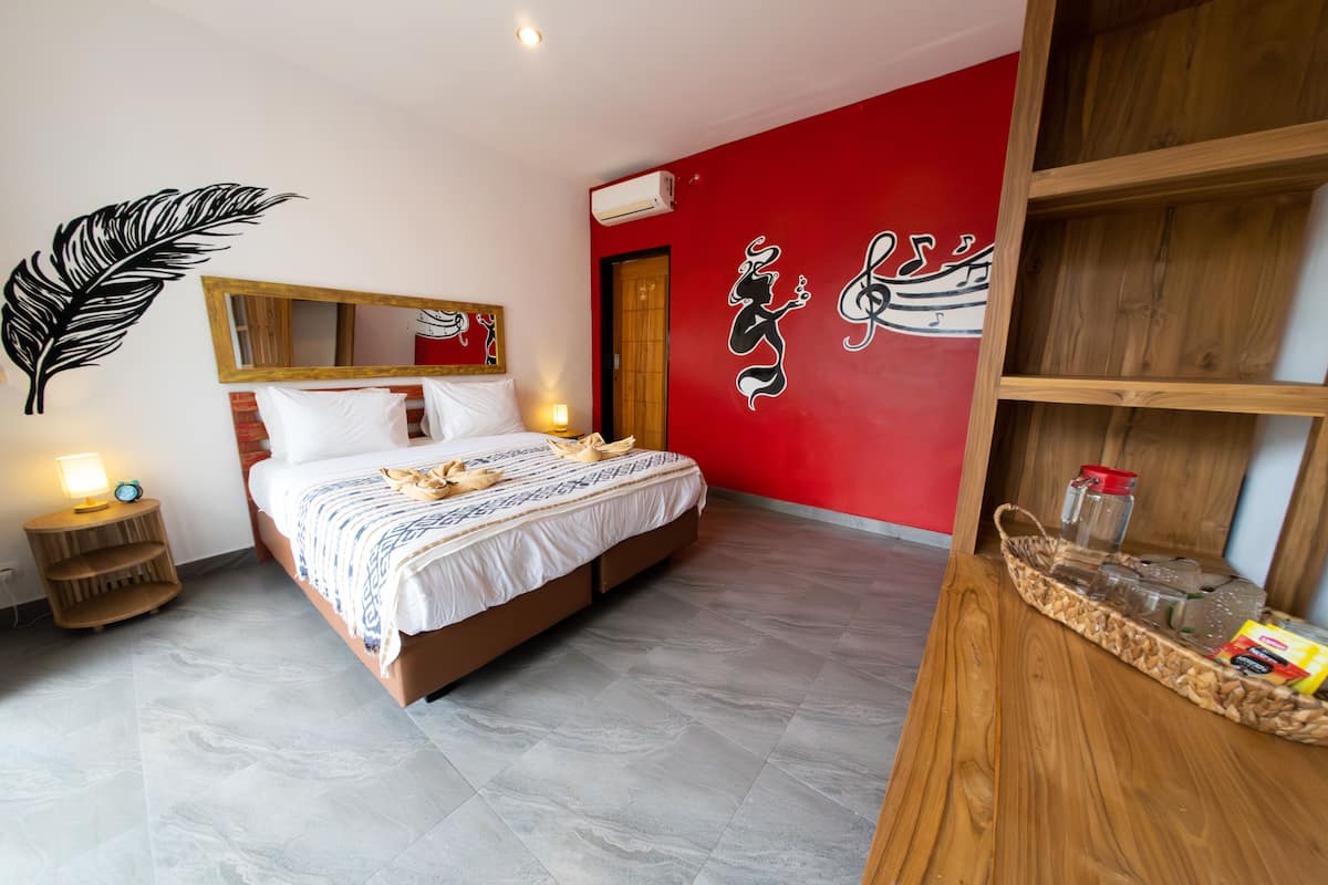 A hotel room with a bed, a nightstand, a mirror, a red wall with decorative art, and a wooden shelf with items.