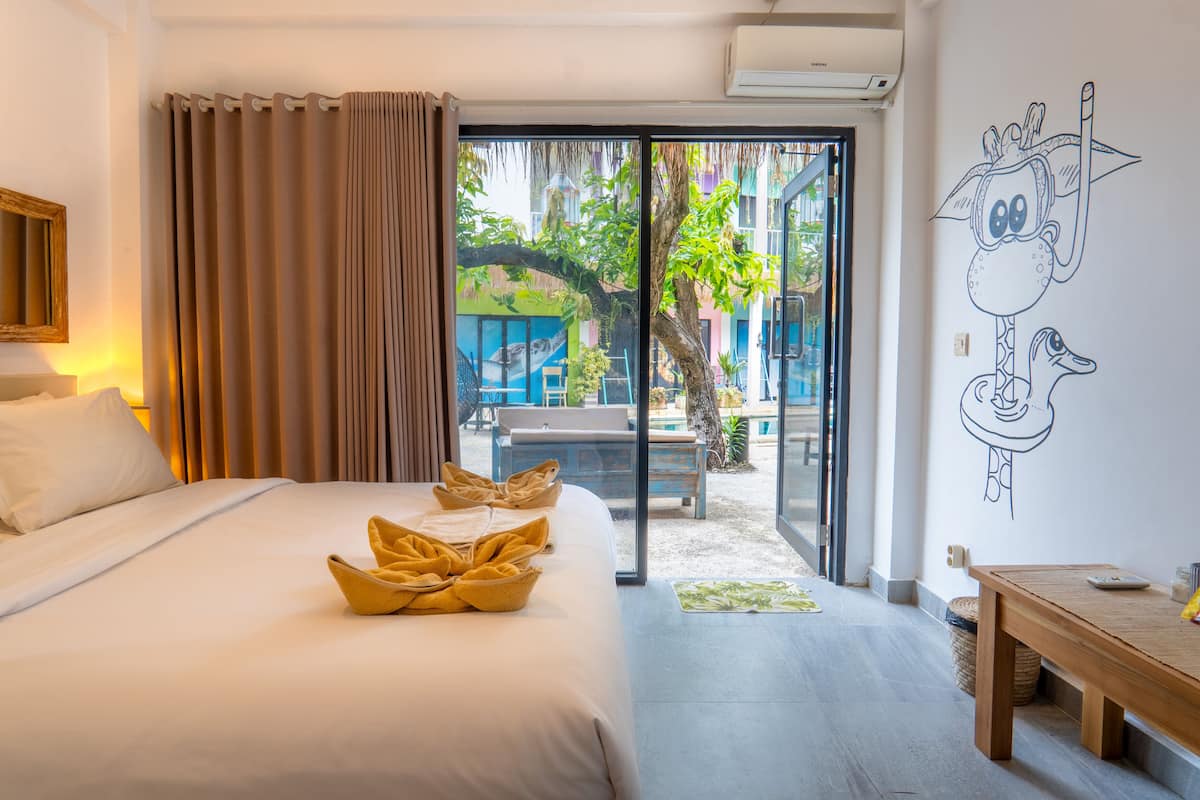 A hotel room with a bed, a wooden bench, a wall mural, and a sliding glass door leading to an outdoor area.