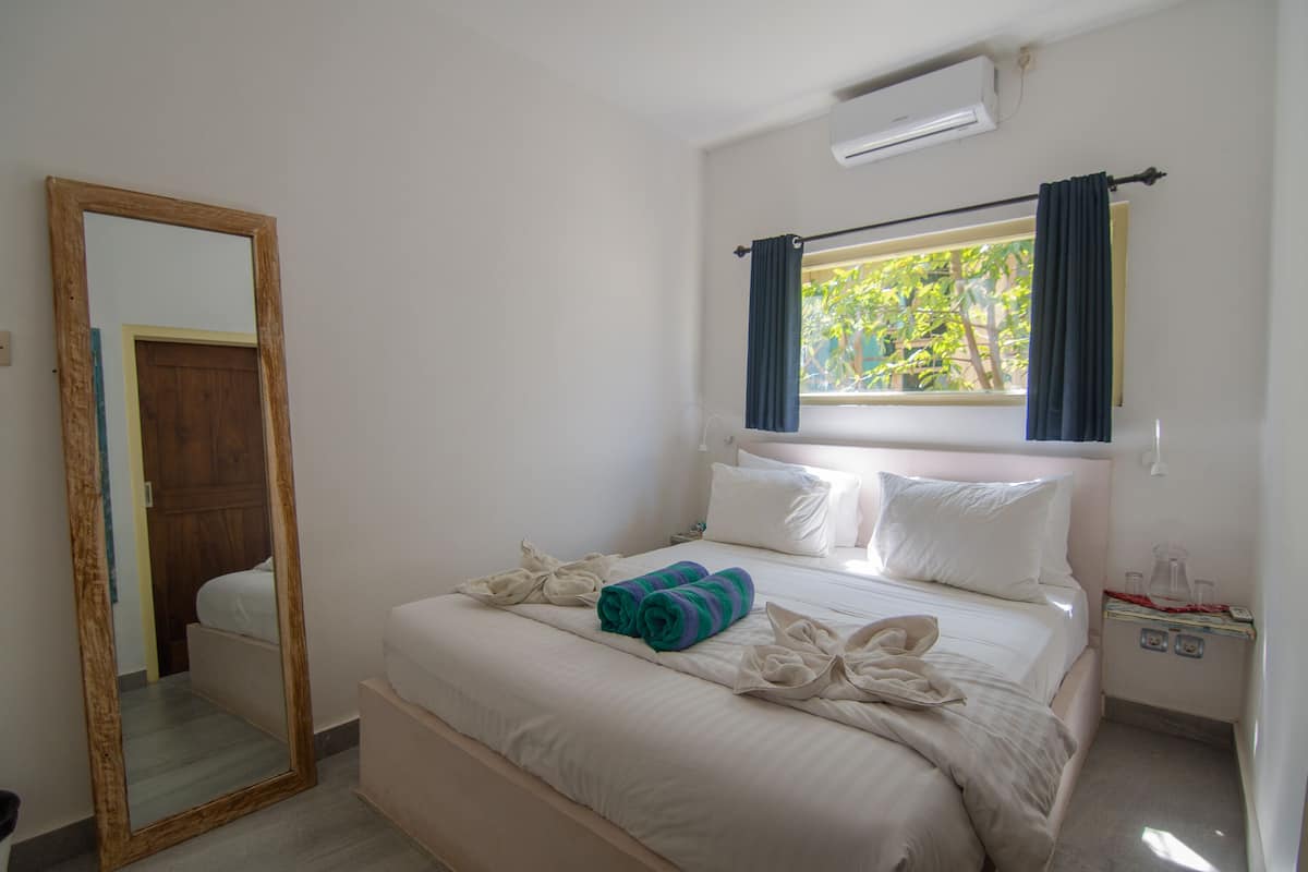 A hotel room with a bed, a large mirror, a window with curtains, and a nightstand with a lamp.