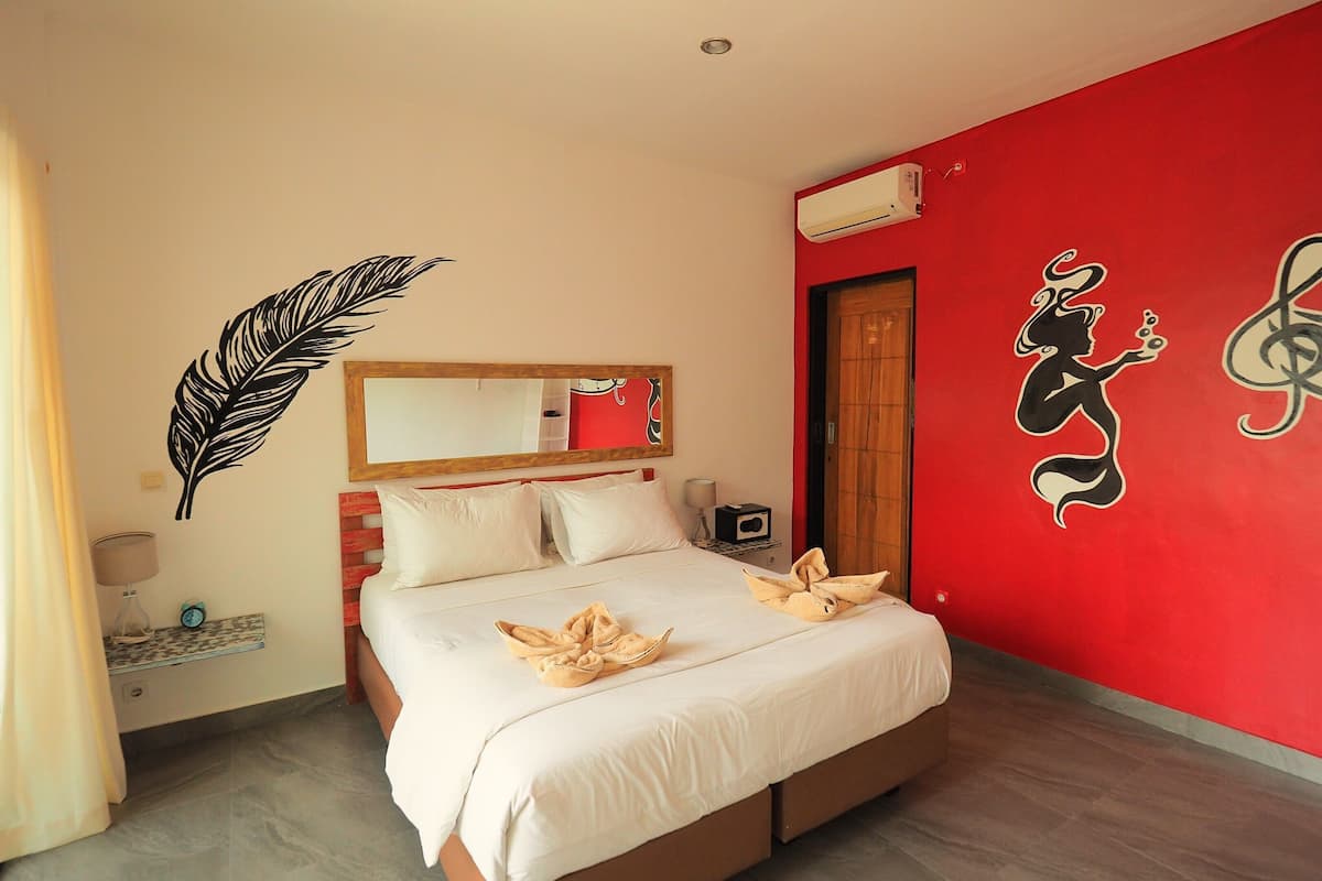 A hotel room with a bed, a mirror, a red wall with a musical note design, a feather decal, and a window with curtains.