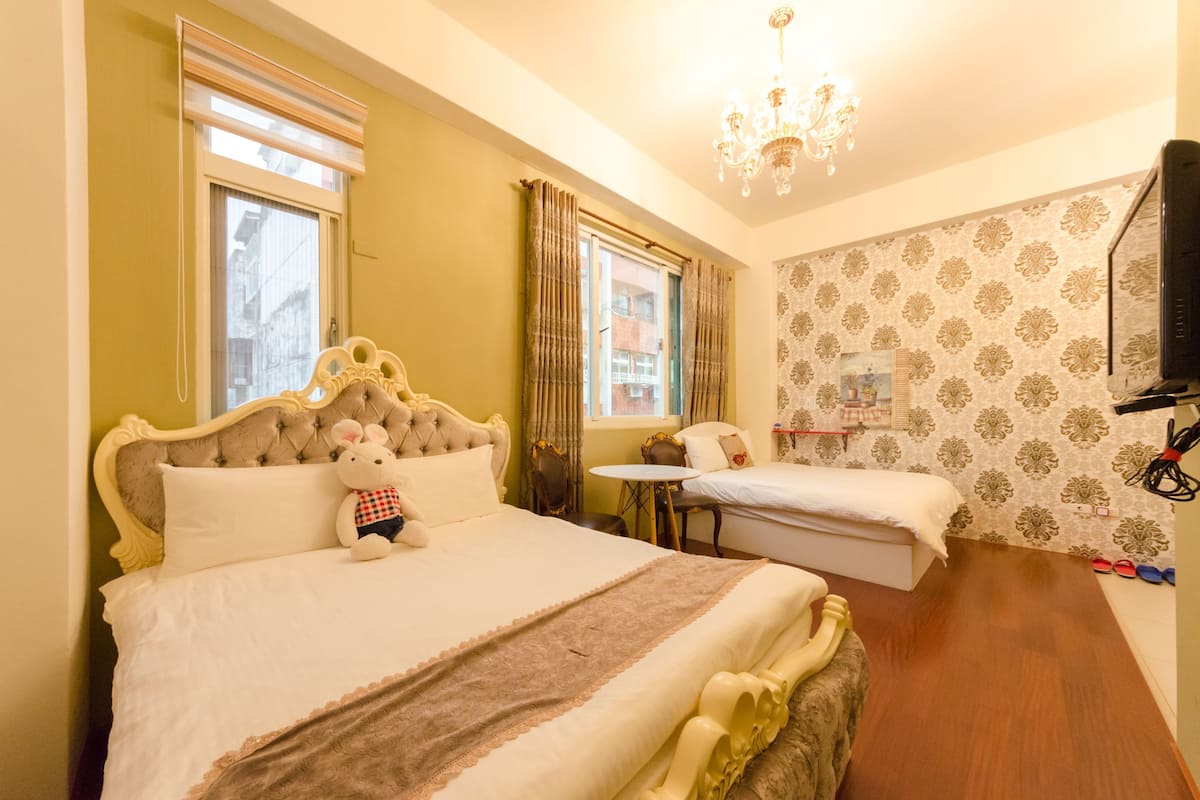 Ein Hotelzimmer mit einem großen Bett, einem kleineren Bett, einem Fernseher, einer Kristallleuchtre und einem Fenster mit Vorhängen.