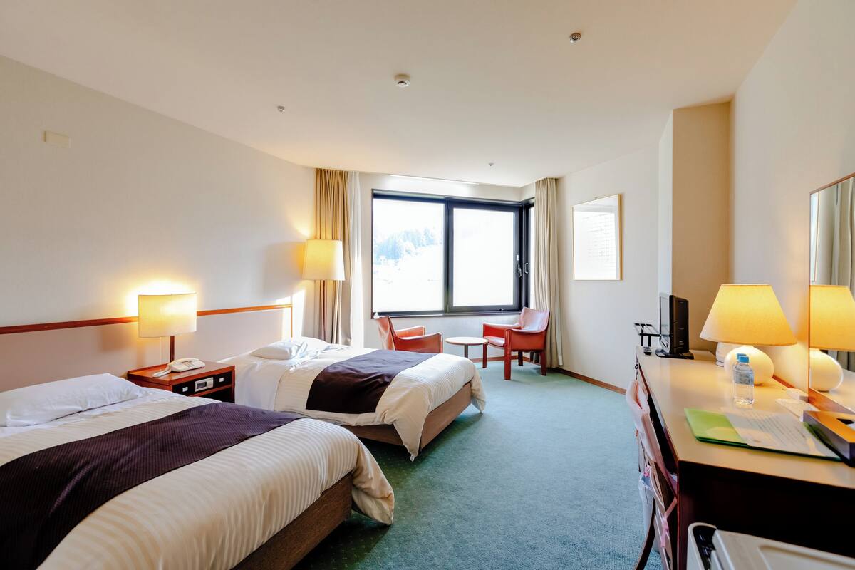 A hotel room with two beds, a desk, a chair, a desk lamp, a telephone, and a window with curtains.