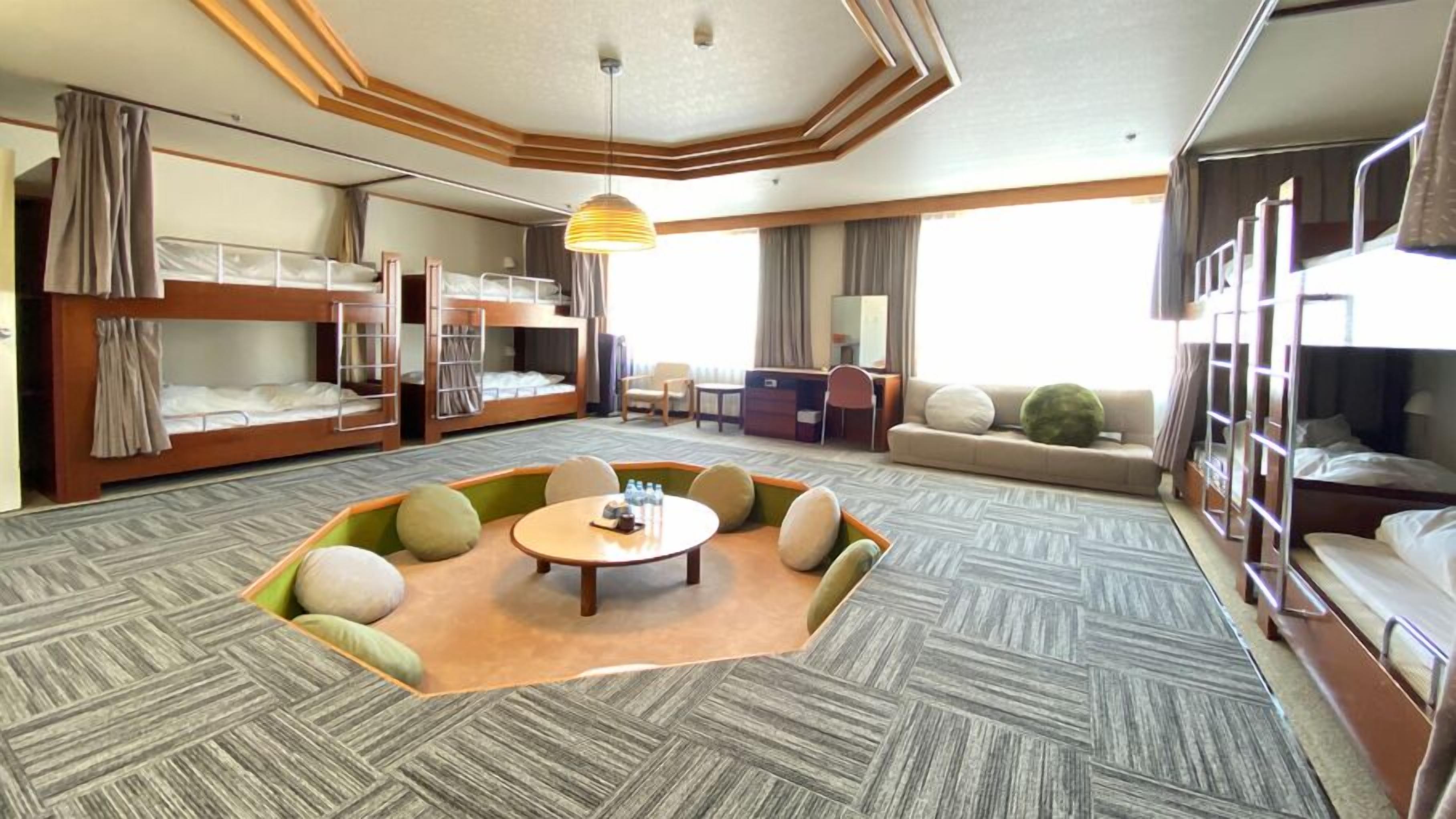 A hotel room with bunk beds, a central seating area, a small table, and a large window.