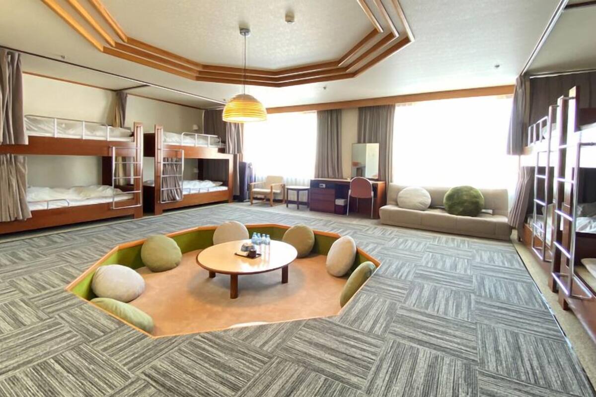A hotel room with bunk beds, a central seating area, a small table, and a large window.