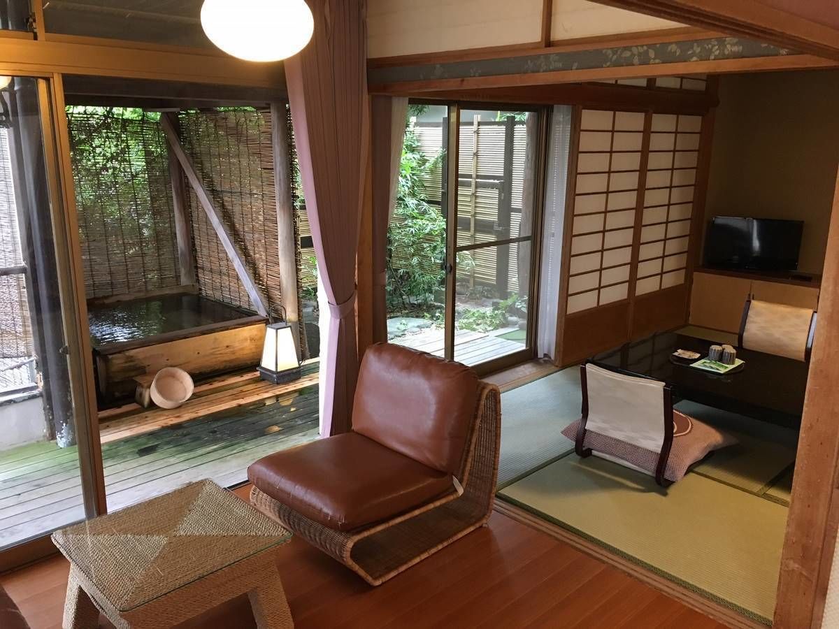 Ein traditioneller japanischer Raum mit Tatami-Bodenbelag, einem braunen Ledersessel, einem niedrigen Tisch mit Fernseher und Blick auf einen Garten durch verschiebbare Türen.