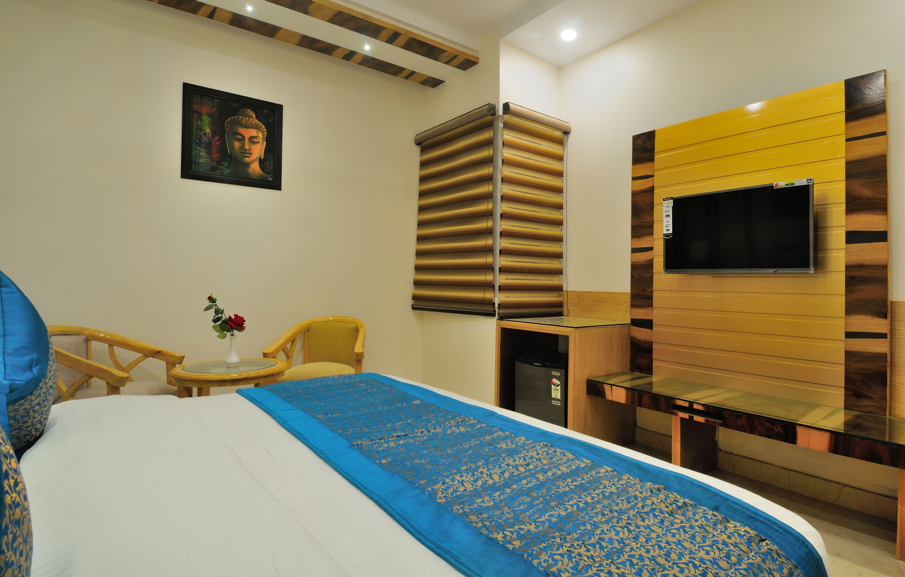Quarto de hotel com cama, televisão na parede, pequena mesa com um vaso de flores e uma cadeira.