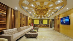 A hotel lobby with a long beige sofa, a reception desk, and a TV screen. - The Gold Regency (New Delhi)