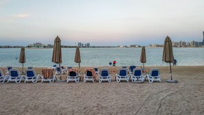 A beach with rows of sun loungers and umbrellas, a calm sea, and a city skyline in the background. - Maison Privee - Charming Apt with Arabesque Sea View on the Palm Jumeirah (Dubai)