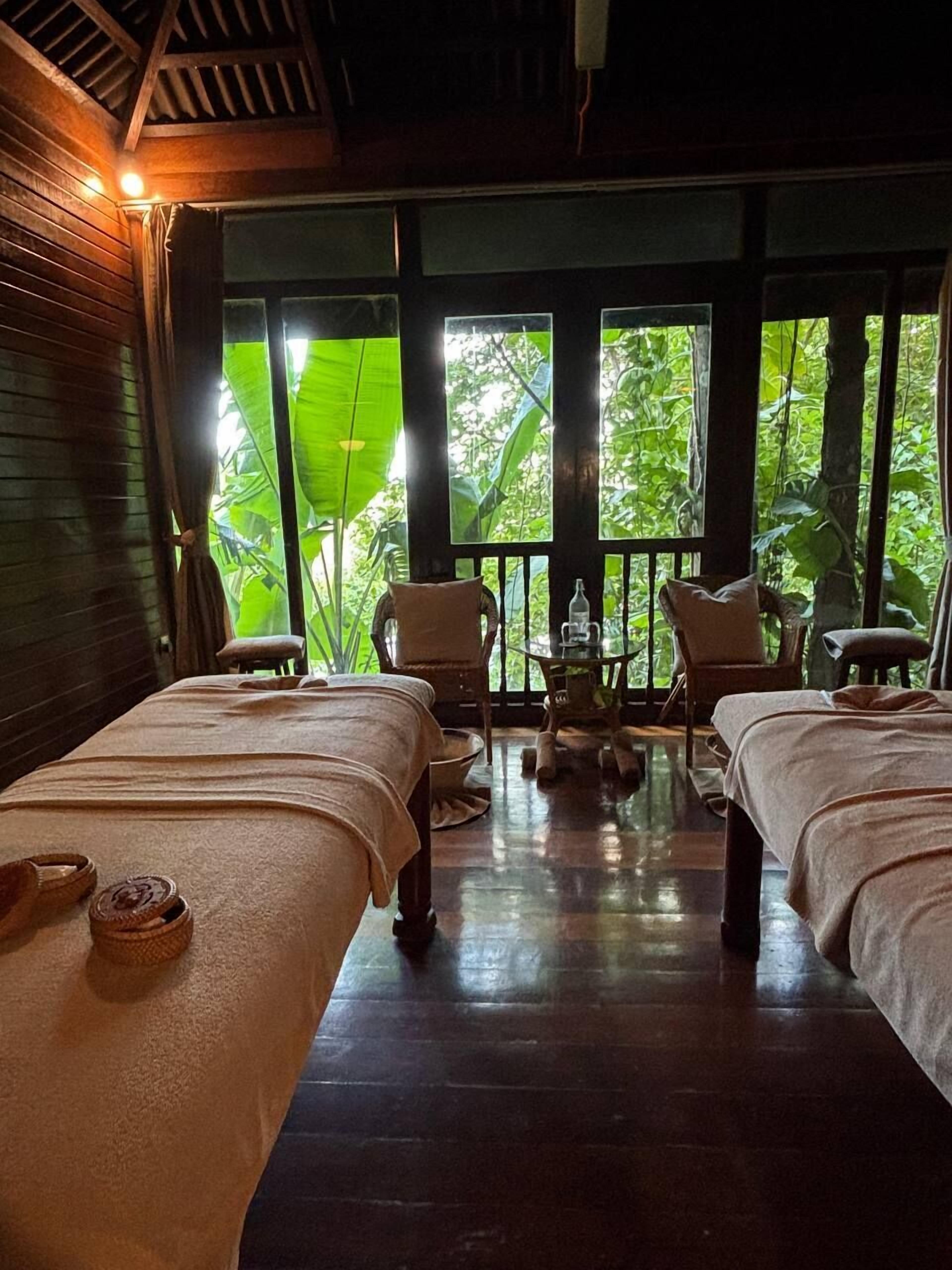 A room with two beds, a small table, chairs, and large windows with a view of greenery.