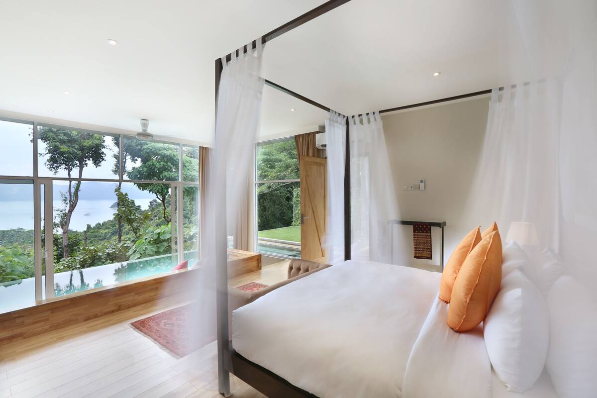 A bedroom with a large bed, white bedding, and orange pillows. It features a large window with a view of greenery and a pool.