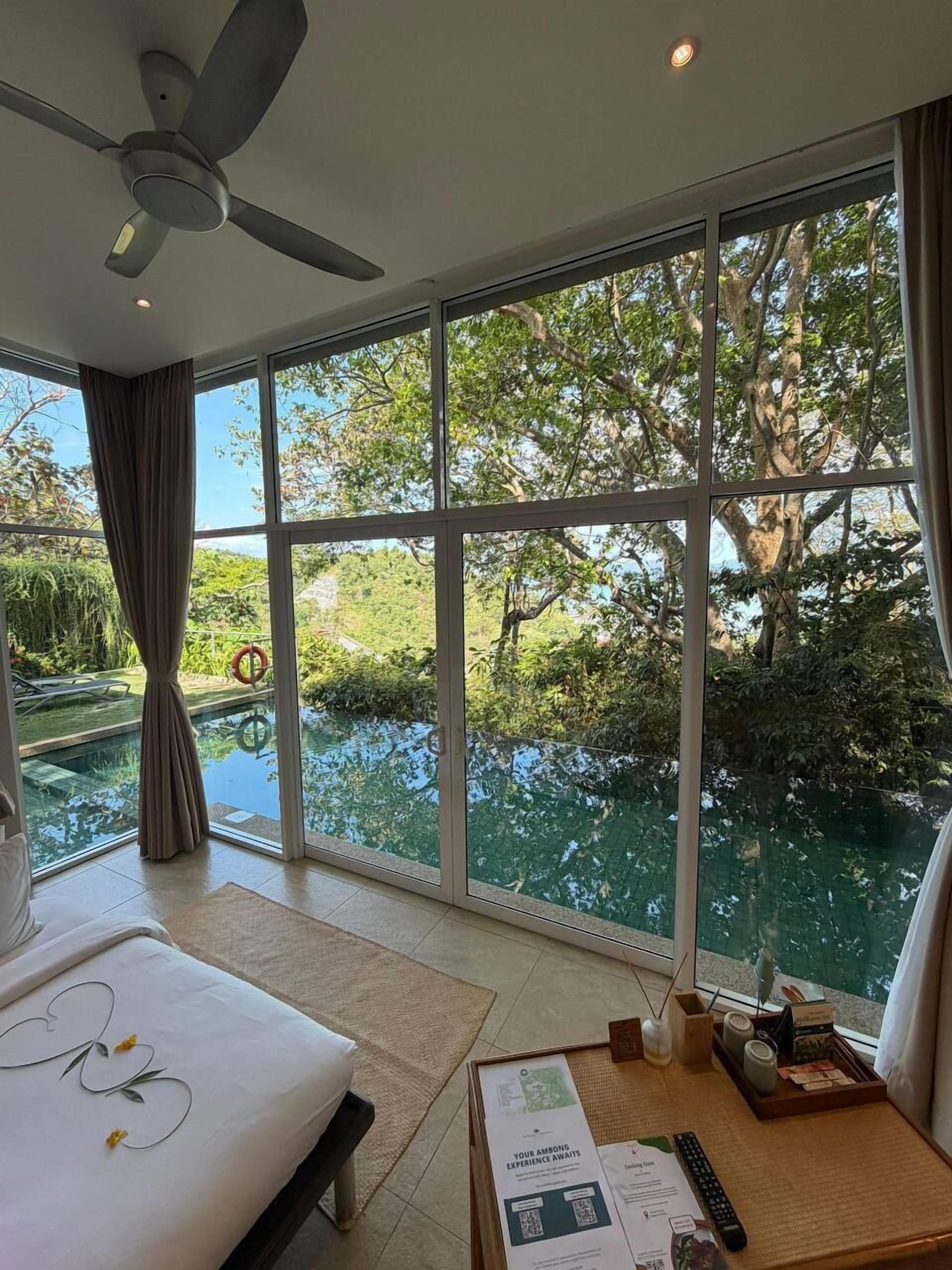 A hotel room with a large window, a ceiling fan, a bed, and a small table with a remote control and a menu.