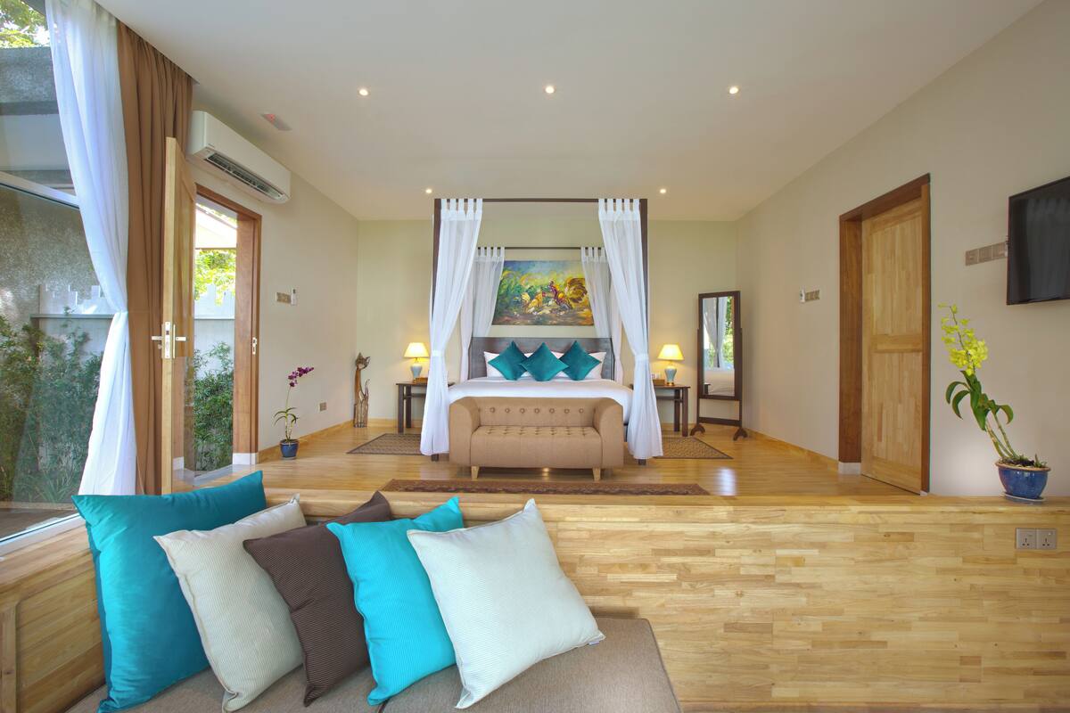 A spacious bedroom with a large bed, a sitting area with cushions, a painting on the wall, and a potted plant.