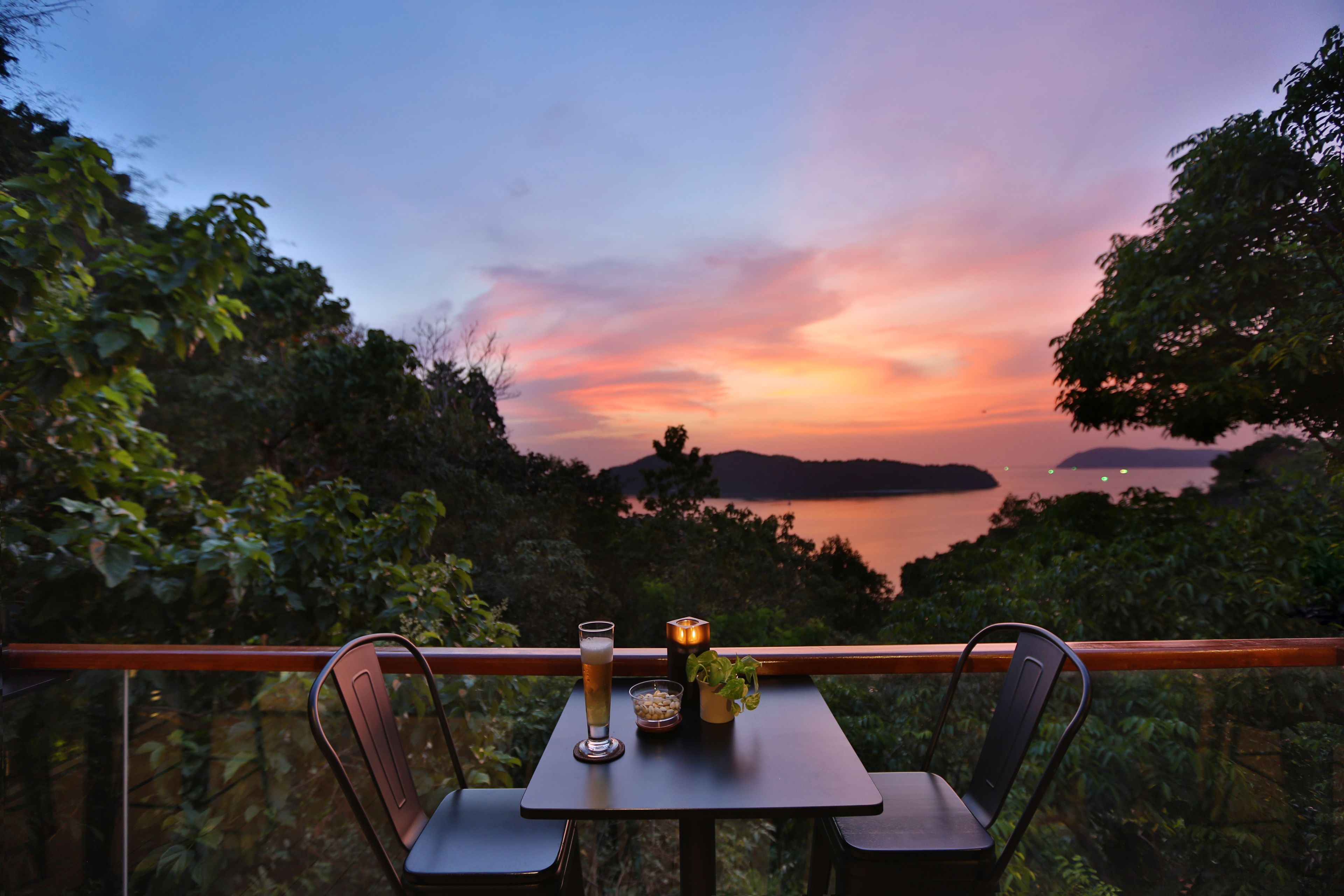 A table with a glass of beer, a candle, and a small plant, overlooking a sunset over a body of water with trees in the foreground.