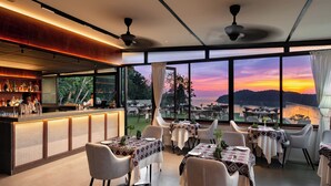 A restaurant with a bar, tables with white chairs, and large windows offering a view of a sunset over a body of water. - Ambong Pool Villas (Langkawi)