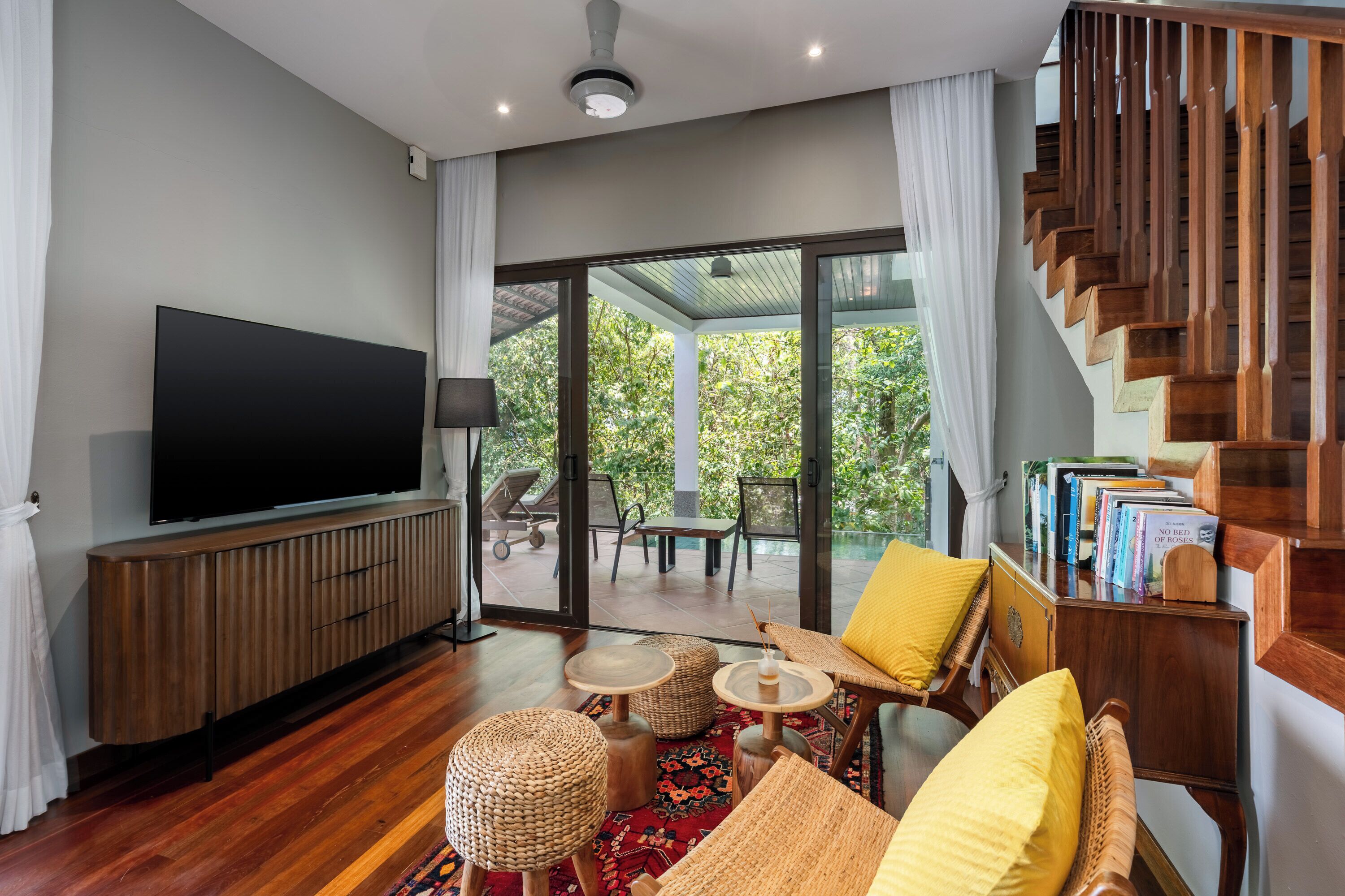 A modern living room with a flat-screen TV, wooden furniture, a staircase, and a view of greenery.