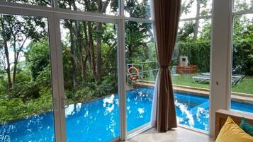 Ett rum med stora fönster med utsikt över en swimmingpool, en takfläkt och en vy över träd och grönområden utanför.