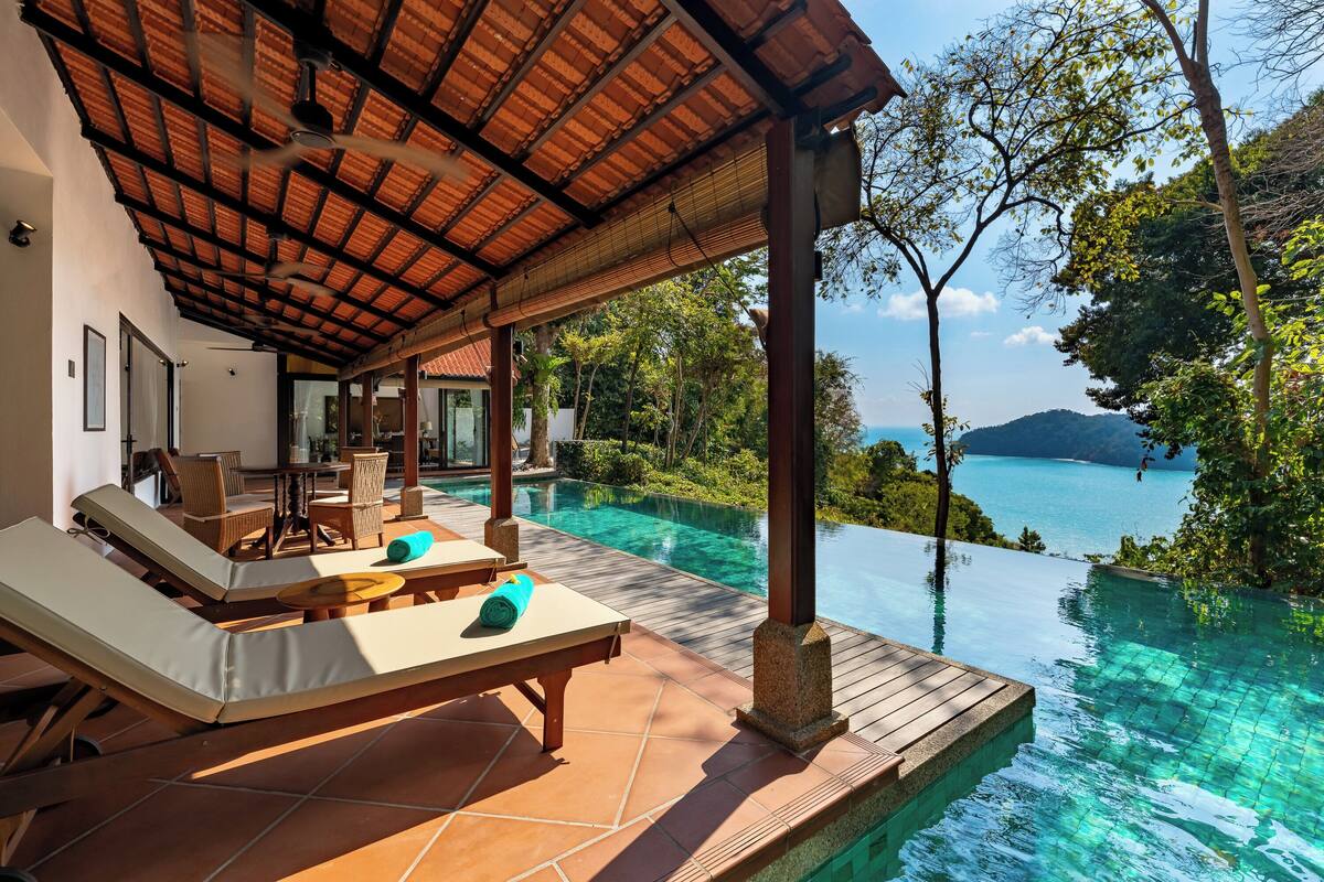 An infinity pool with clear water, a wooden deck, lounge chairs, and a covered seating area with a view of the sea.