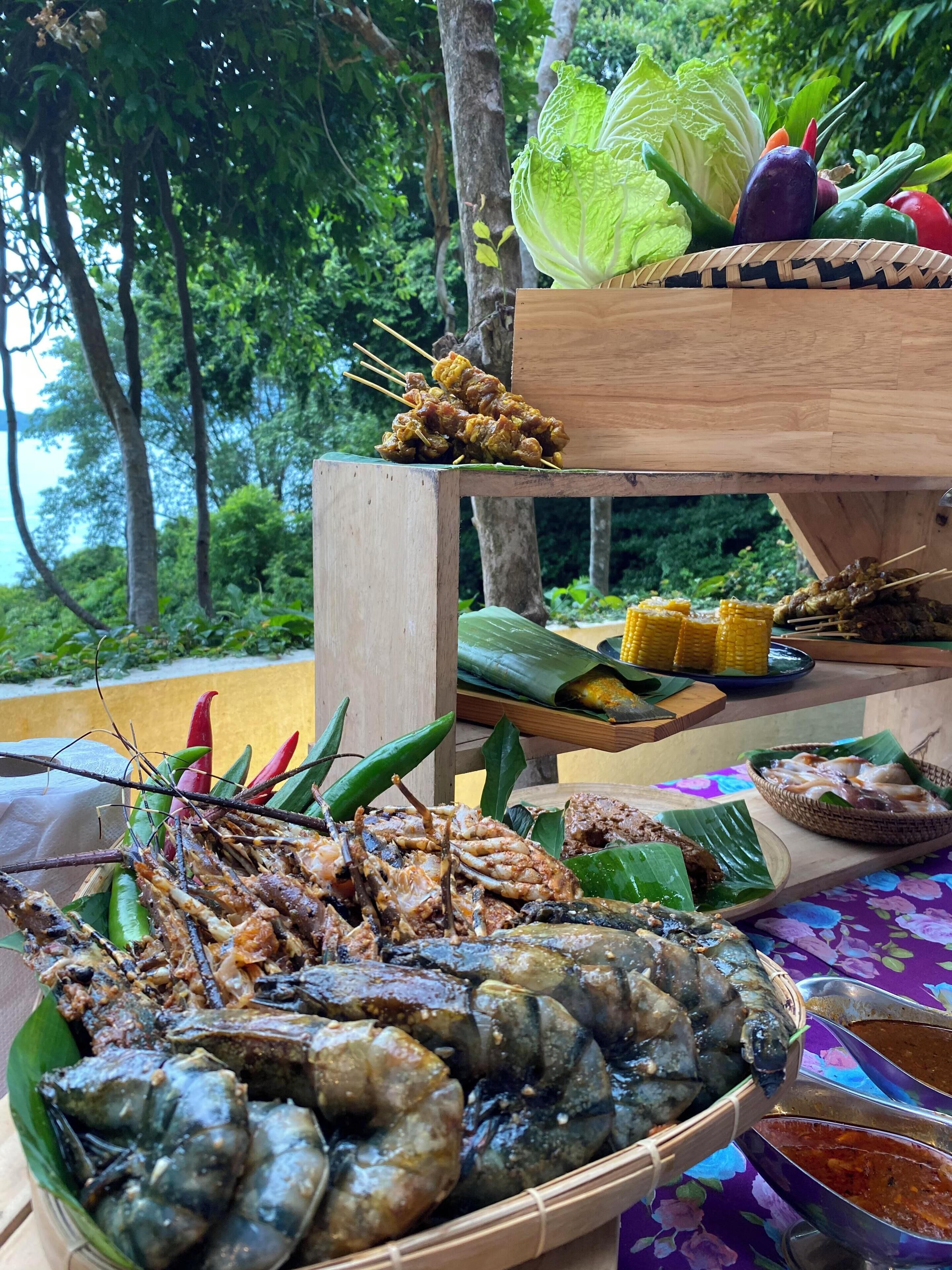 Diversas preparaciones a la parrilla de mariscos y verduras sobre una mesa de madera al aire libre, con un exuberante fondo verde.