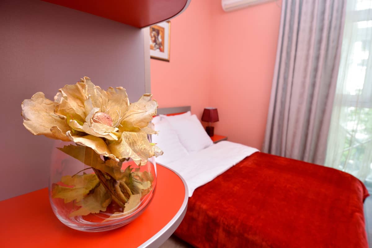A hotel room with a bed, a red bedside table, and a vase with dried flowers.