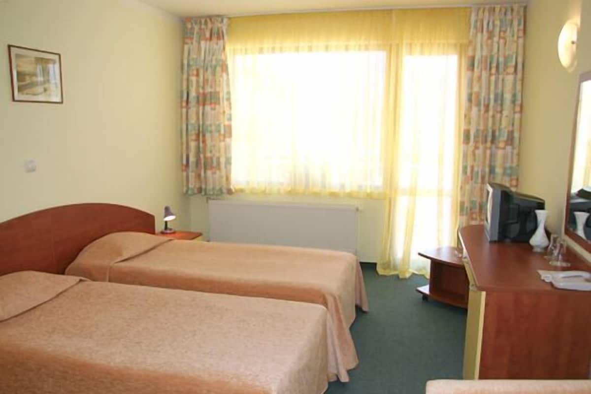 A hotel room with two beds, a desk, a chair, a radiator, and a window with curtains.